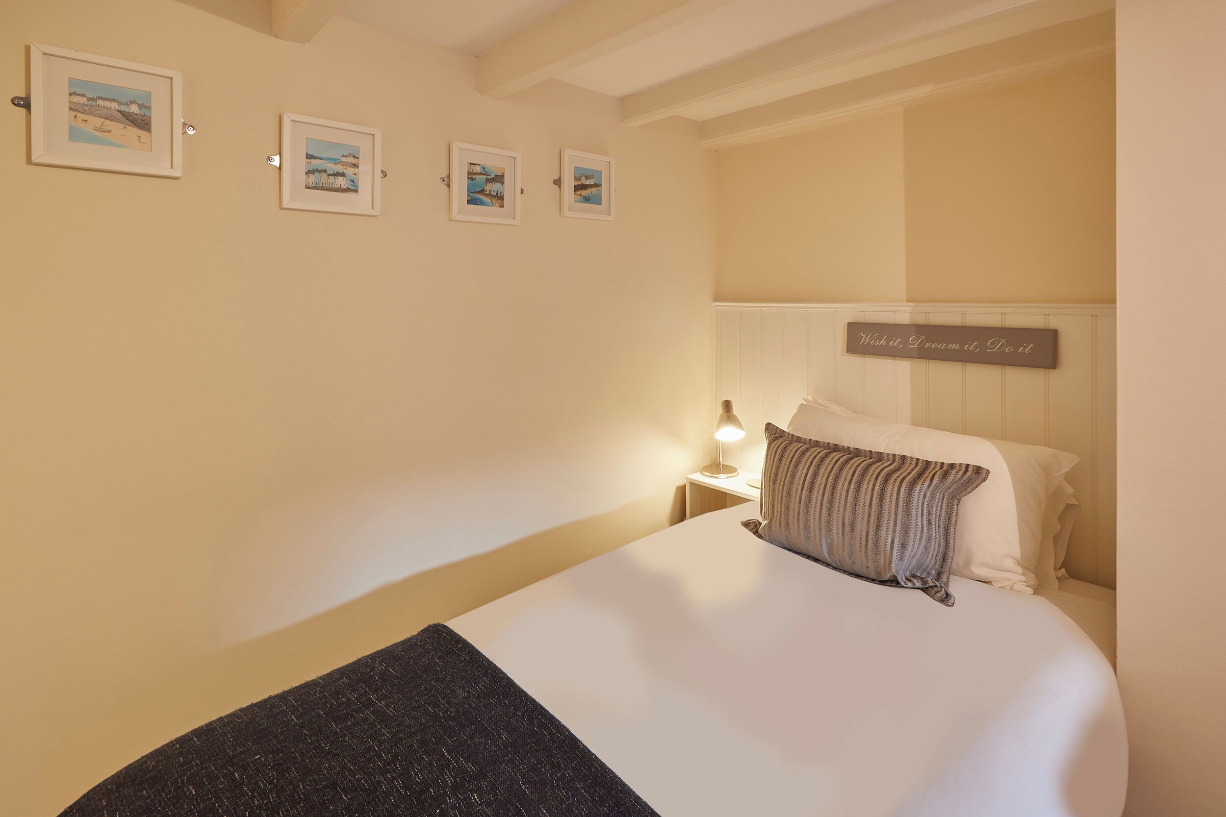 A neatly made bed with a pillow and a blanket, a bedside lamp, and a wall-mounted sign with the text "Wish it, Dream it, Do it".