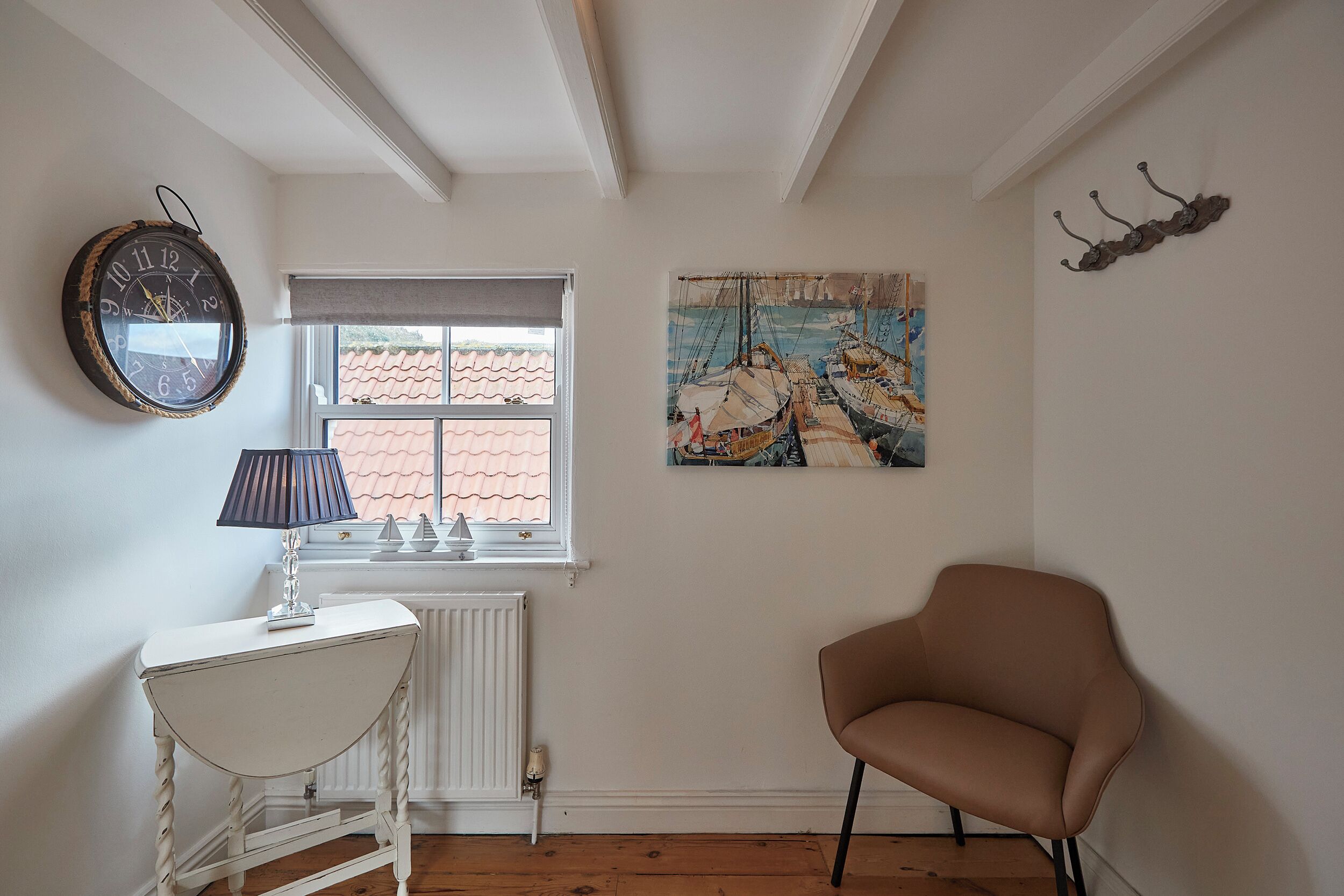 A room with a white radiator, a round wall clock, a window with a view of a tiled roof, a brown chair, and a painting of sailing ships.