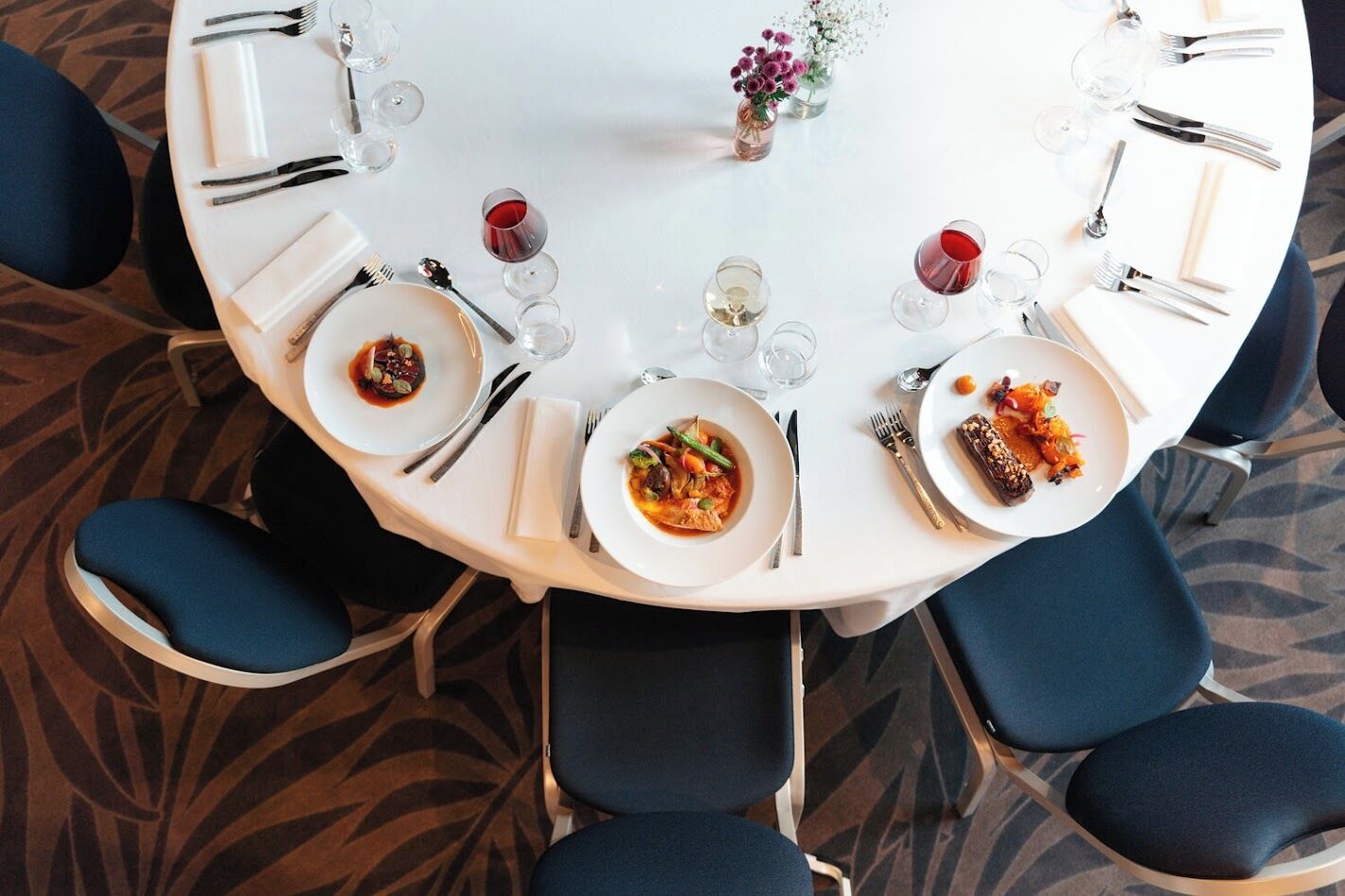 A round dining table with white plates, cutlery, and wine glasses, surrounded by blue chairs.
