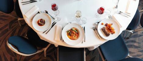 A round dining table with white plates, cutlery, and wine glasses, surrounded by blue chairs.