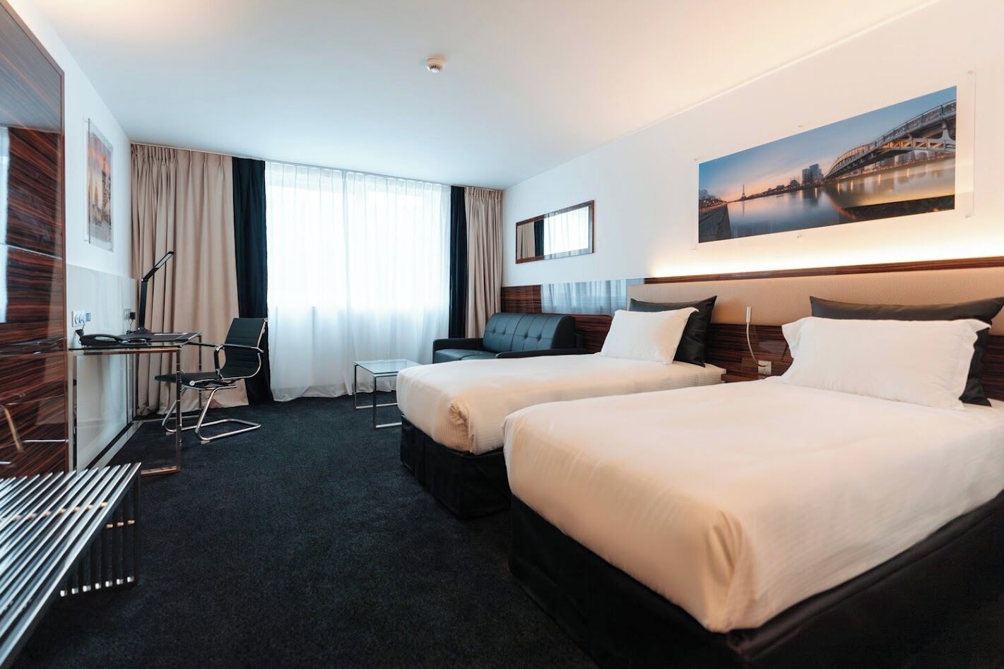 A hotel room with two beds, a desk, a chair, and a large window with curtains.