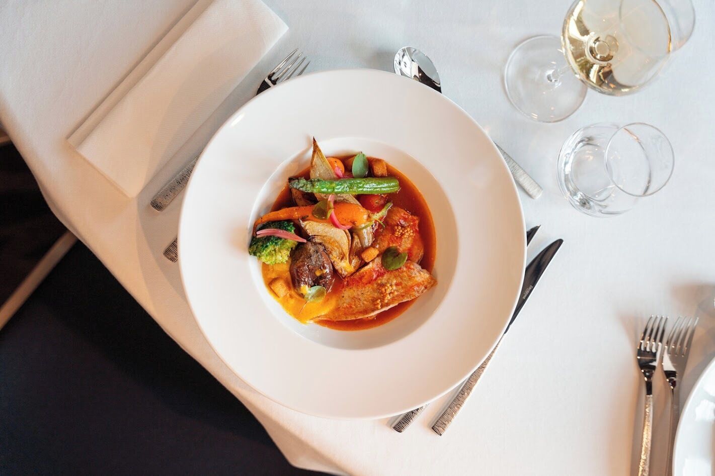 Une assiette blanche contenant un ragoût de légumes et de viande, accompagnée d’une fourchette et d’un couteau posés sur une nappe blanche.