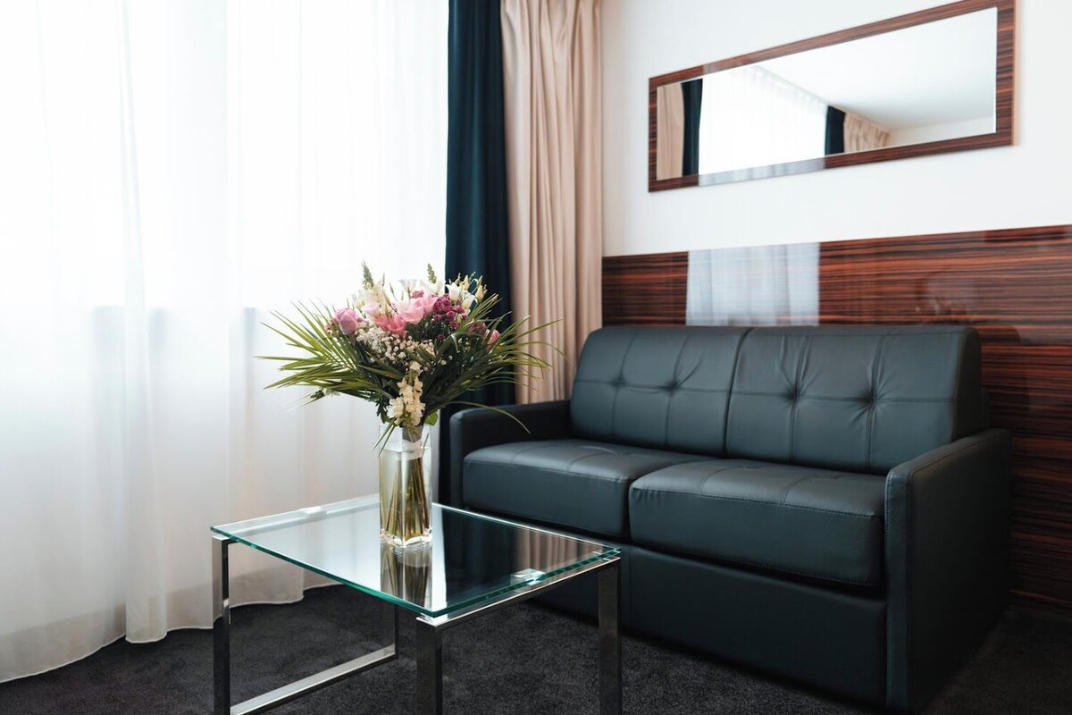 A modern hotel room with a glass coffee table, a dark leather sofa, and a large mirror on the wall.