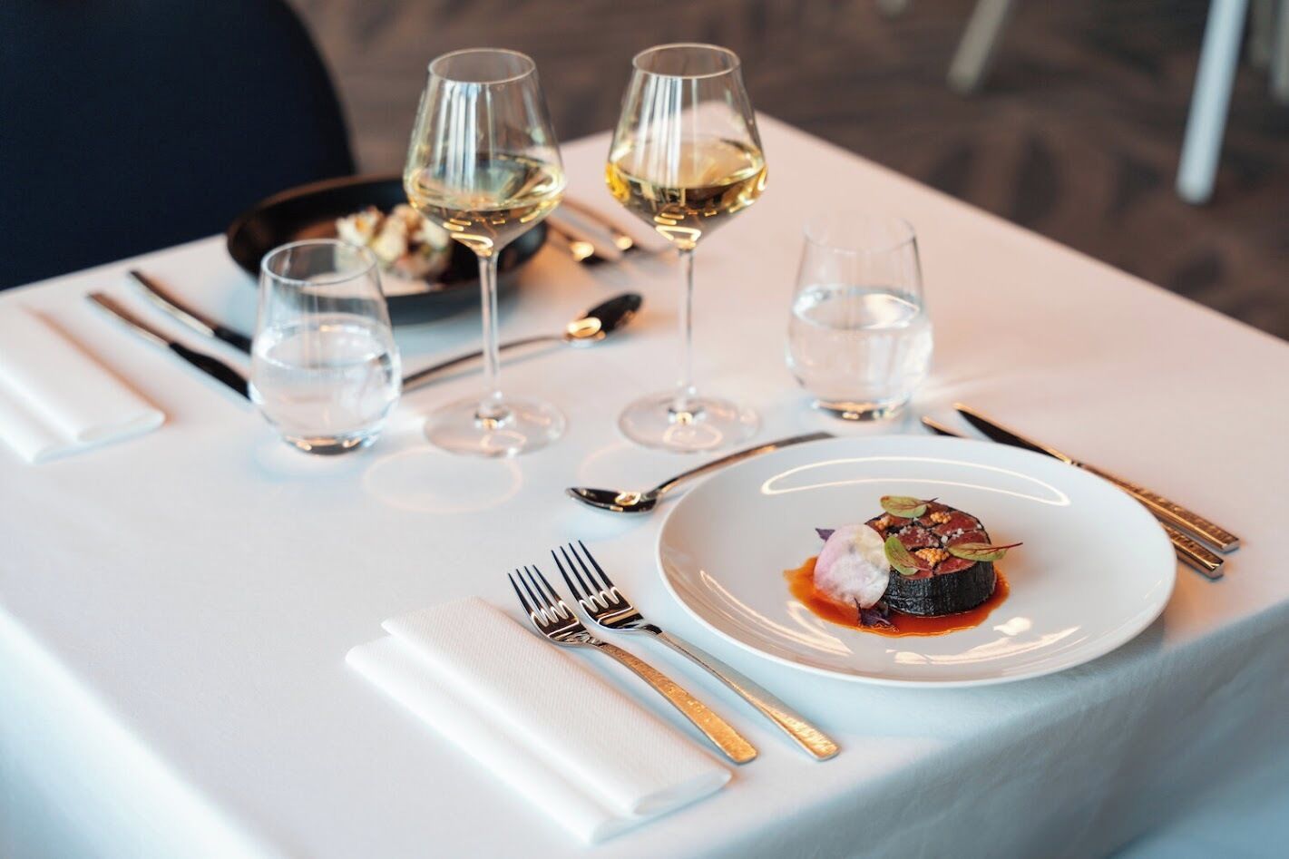 Una mesa dispuesta para una comida, con mantel blanco, copas de vino, cubiertos y un plato de comida.