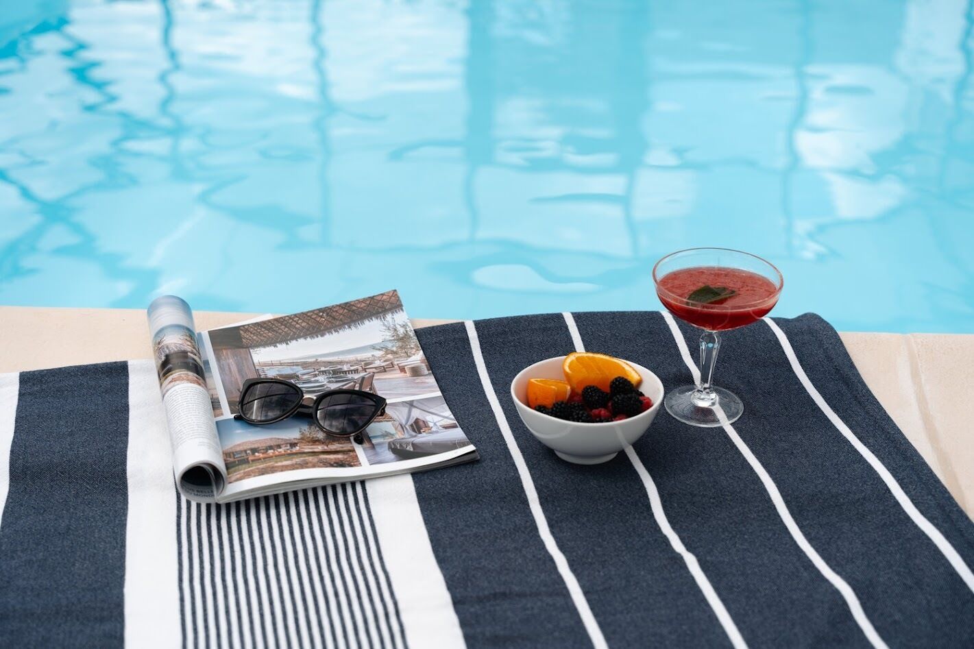 A poolside setting with a magazine, sunglasses, a bowl of fruit, and a glass of red drink on a striped towel.