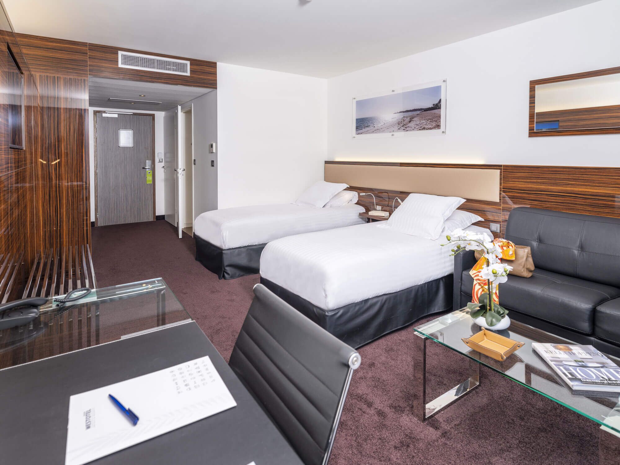 Ein Hotelzimmer mit zwei Betten, einem Schlafsofa, einem Glastisch und einem Schreibtisch mit Stuhl.