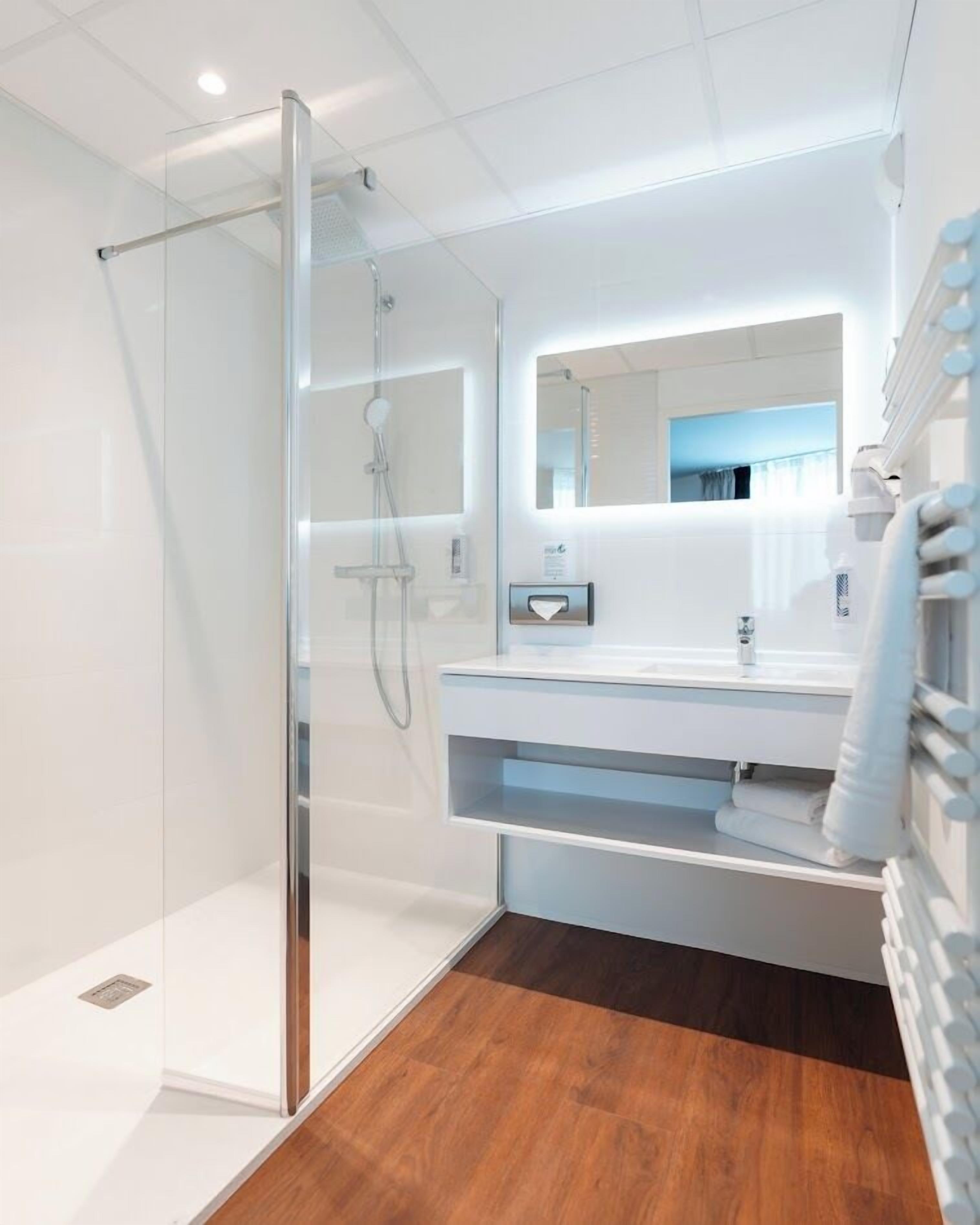 A modern bathroom with a glass shower enclosure, a white vanity with a mirror, and a wooden floor.