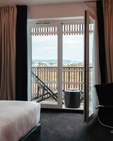 Habitación de hotel con cama, una silla, una mesita y balcón con vistas a la ciudad.