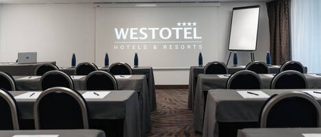 Un salón de conferencias en Westotel Hotels & Resorts con filas de mesas, sillas, una pantalla de proyector y una computadora portátil sobre una mesa.