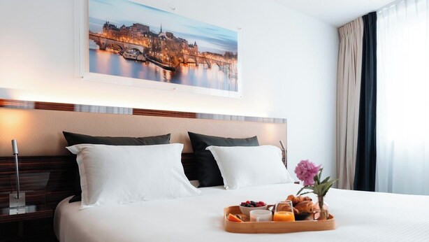 A hotel room with a large bed, two pillows, a tray with breakfast items, a nightstand, a lamp, and a painting of a cityscape above the bed.
