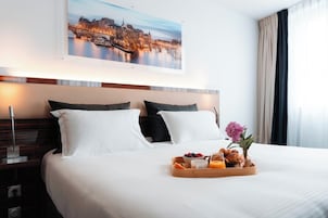 Habitación de hotel con una cama grande, dos almohadas, una bandeja con artículos de desayuno, una mesita de noche, una lámpara y un cuadro de un paisaje urbano sobre la cama.