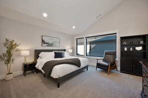 Une chambre moderne avec un grand lit, une table de chevet, une lampe, une chaise, une commode et une fenêtre avec des stores.