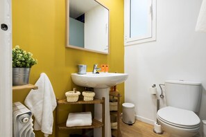 Une salle de bain avec un mur jaune, un lavabo blanc et des toilettes. On y trouve des paniers sur une étagère, une serviette suspendue et une poubelle.