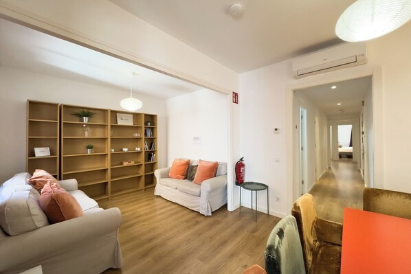 Un salon moderne avec un canapé, un fauteuil blanc et une bibliothèque en bois. On y trouve une petite table sur laquelle est posé un objet rouge, ainsi qu’un extincteur fixé au mur.