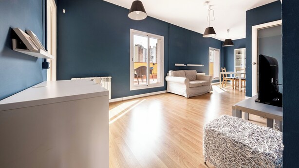 Ein modernes Wohnzimmer mit einem weißen Sofa, einem Couchtisch und einem Fernseher. Es gibt zwei Pendelleuchten, einen weißen Hocker und ein Bücherregal.
