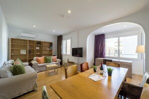Una sala de estar moderna con sofá, mesa de comedor y televisor. Hay estanterías y un ventanal con cortinas.