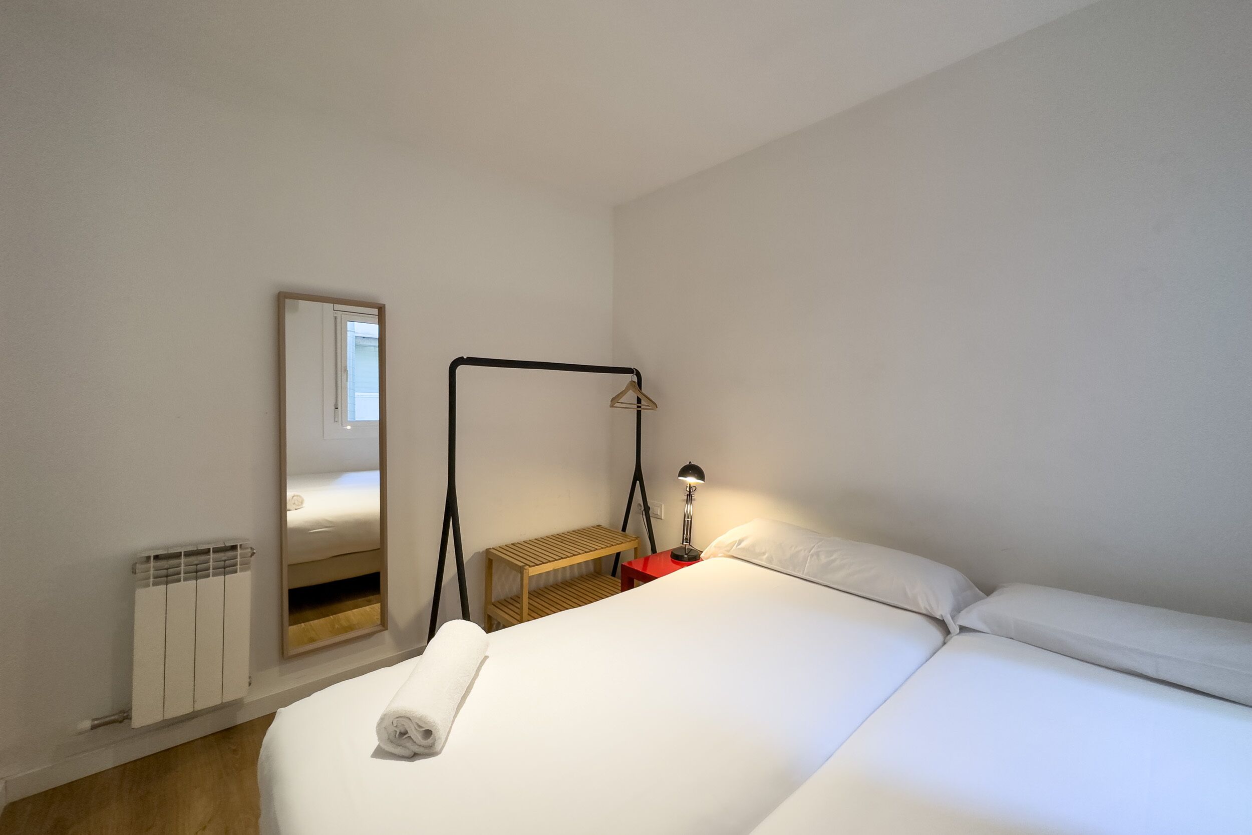 A neatly made bed with white linens, a bedside table with a lamp, a mirror, and a radiator.