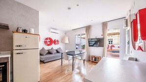 Una cocina moderna con un refrigerador blanco, un comedor con mesa y sillas de vidrio, un sofá gris y un televisor en la sala.