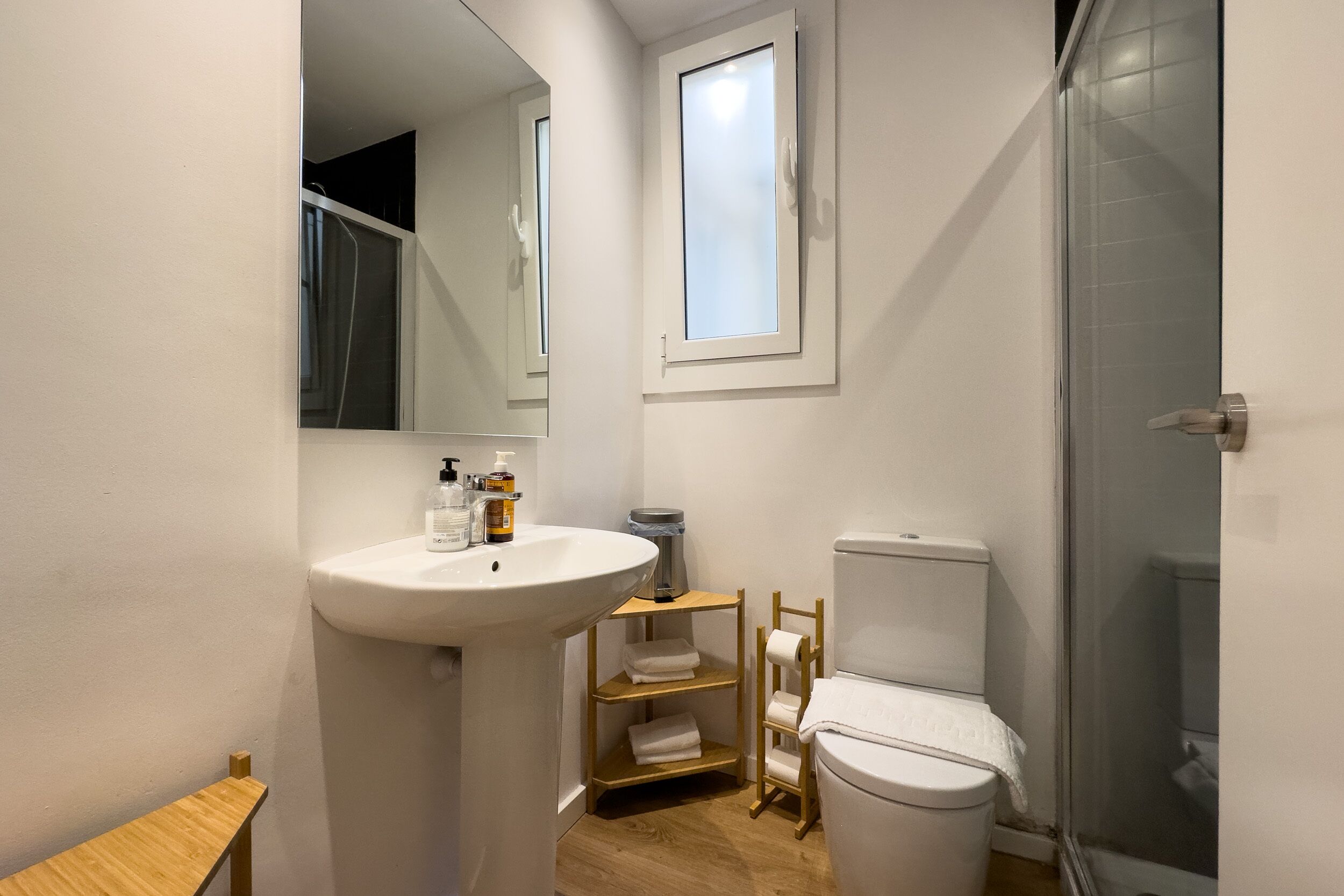 A compact bathroom with a white sink, toilet, and shower. There is a wooden shelf with towels and toiletries.