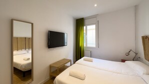 Ein Hotelzimmer mit zwei Betten, einem Fernseher, einem kleinen Holztisch und einem Fenster mit grünen Vorhängen.