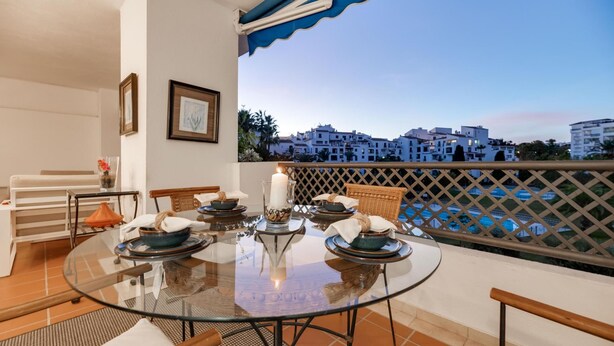 A dining area with a glass table, chairs, and a view of buildings and a pool. - Urban Exclusivity At Puerto Banús (Marbella)