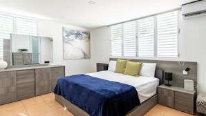 A modern bedroom with a large bed, a nightstand, a bedside table, a mirror, and a window with blinds. - Luxury Casa CALIAD (San Juan)