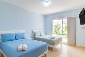Quarto de hotel com duas camas, televisão e uma janela grande com vista para palmeiras.