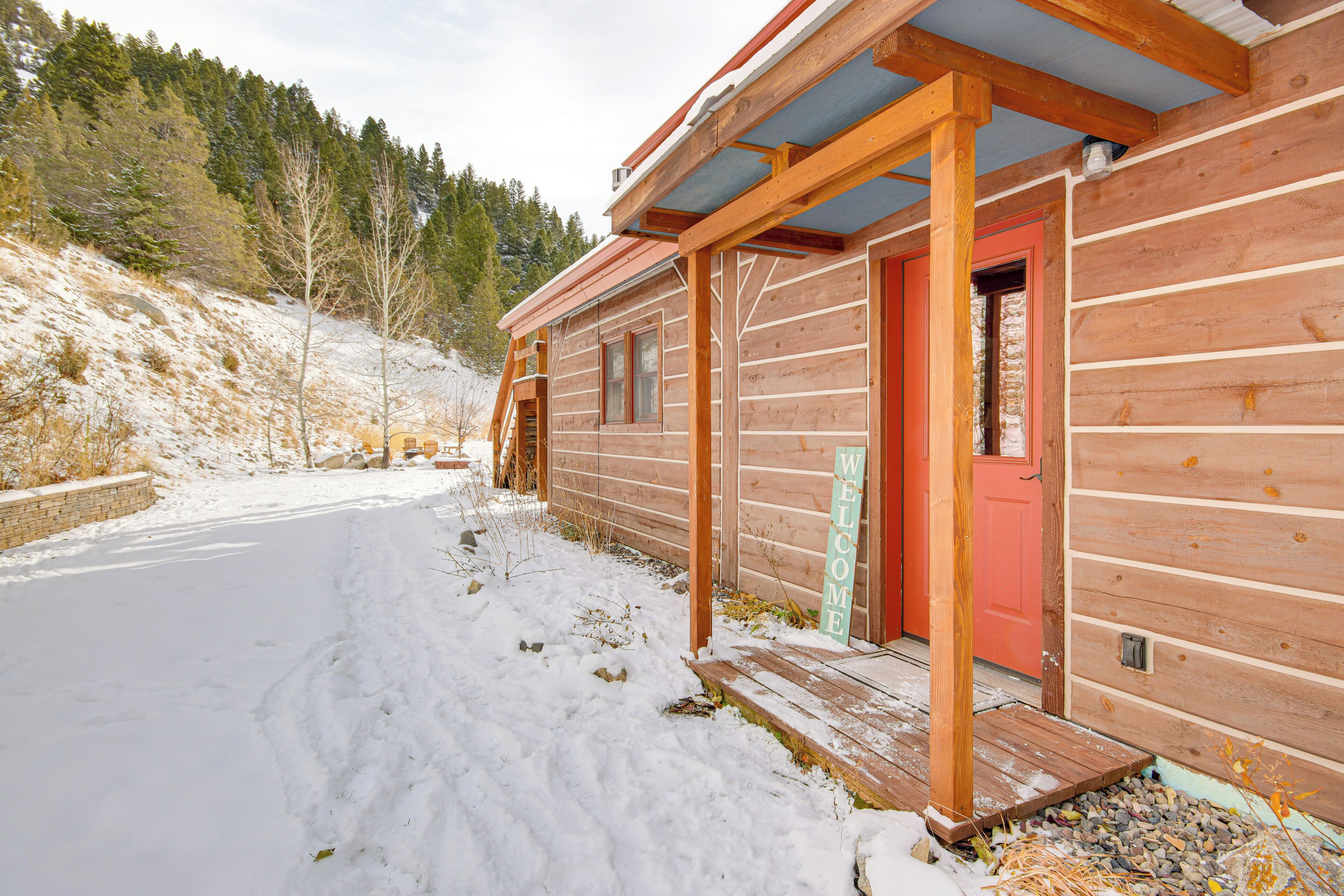 Una cabaña de madera con puerta roja y un cartel que dice 'WELCOME', rodeada de nieve y un paisaje boscoso.