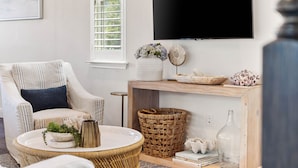 Una sala de estar con un sillón a rayas, una mesa redonda de mimbre, una consola de madera con objetos decorativos y un televisor montado.