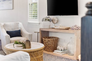A living room with a striped armchair, a round wicker table, a wooden console table with decorative items, and a mounted TV.