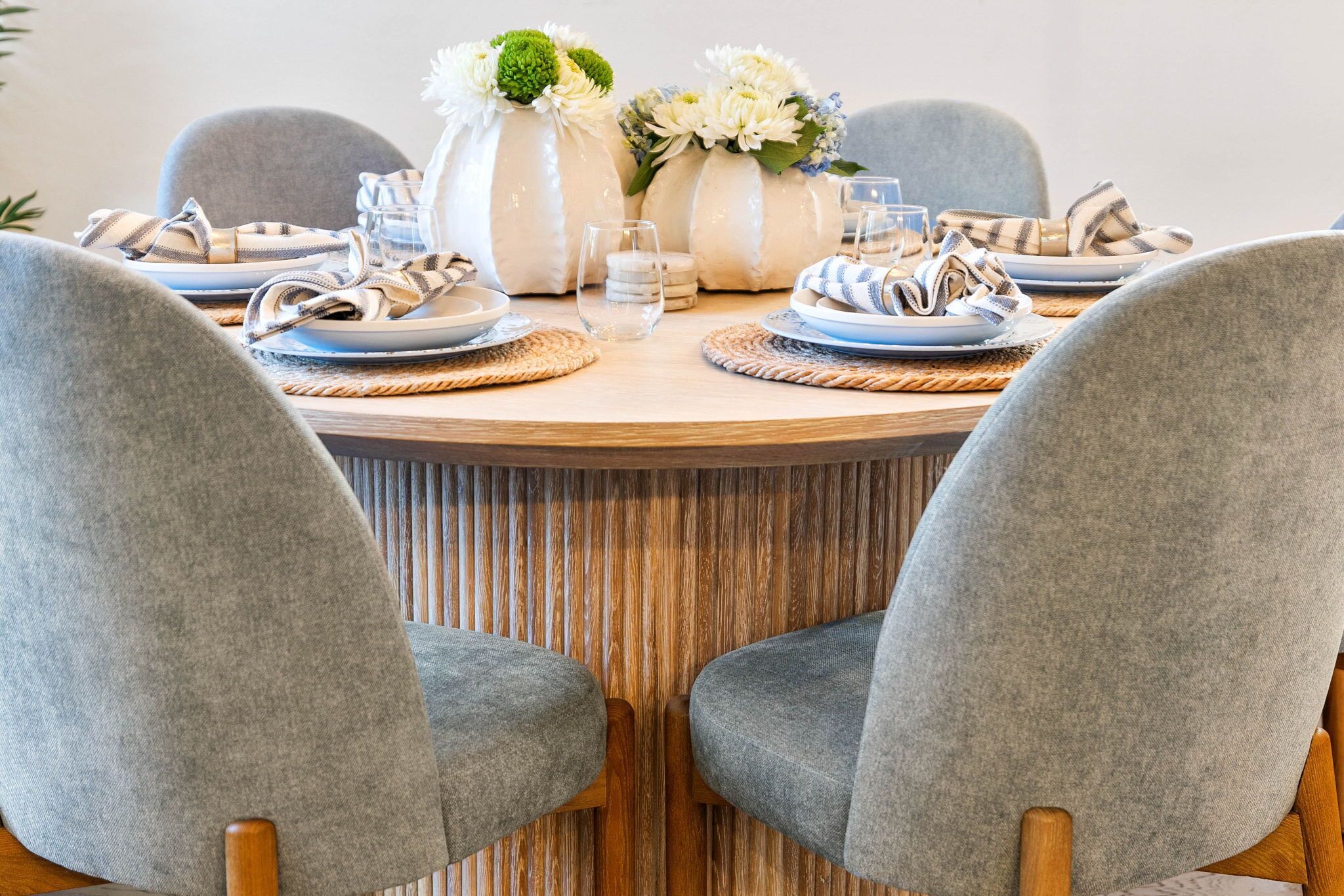 Una mesa redonda para comer con sillas tapizadas en gris, platos blancos y azules, y servilletas a rayas. Hay jarrones con flores blancas y cristalería transparente sobre la mesa.