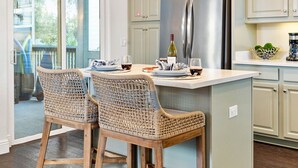 Une cuisine dotée d’un îlot central, de deux chaises en osier, d’un réfrigérateur en acier inoxydable et de luminaires suspendus.