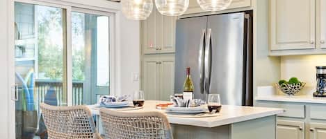 Una cocina con una isla central, dos sillas de mimbre, un refrigerador de acero inoxidable y luces colgantes.