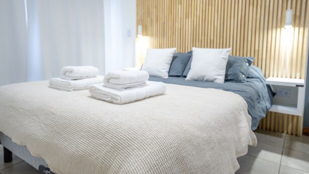 Una cama bien tendida con sábanas y almohadas blancas, una mesita de noche con lámpara, y un cabecero de listones de madera.