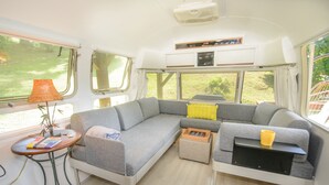 En hyggelig autocamper indretning med en krogsofa, et lille bord med en lampe og et vindue med udsigt til grøn vegetation.