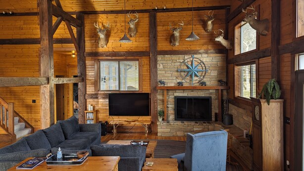 Een rustiek blokhutinterieur met houten wanden, een stenen open haard, opgeplakte hertenkoppen, een flatscreen televisie en een gezellige zithoek met een bank en fauteuils.