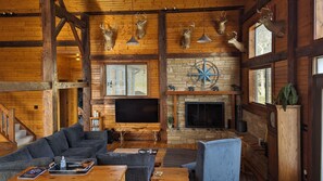 A rustic cabin interior with wooden walls, a stone fireplace, mounted deer heads, a flat-screen TV, and a cozy seating area with a sofa and armchairs.