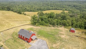 An aerial view of a rural property with two red barns, a large open field, and a forested hill in the background. - The Little Red Schoolhouse (Cross Junction)