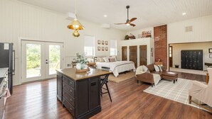A spacious living area with a large wooden dining table, a sofa, and a coffee table. There is a bed with a headboard and a ceiling fan. The room has wooden floors and white walls. - The Little Red Schoolhouse (Cross Junction)