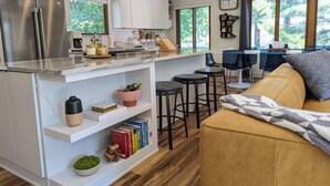 Una cocina moderna con un refrigerador de acero inoxidable, una estufa integrada y un amplio ventanal. Hay una zona de estar con un sofá y una mesa de centro.