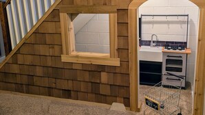 Una casita de juegos con fachada de madera, una ventana, una pequeña cocina con fregadero y estufa, y un carrito de compras de juguete.
