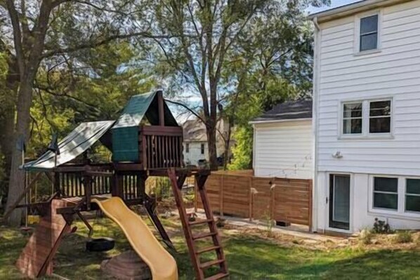 A backyard with a wooden play structure including a slide and climbing area, adjacent to a white house with a garage.