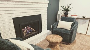 A living room with a fireplace, a dark grey armchair, a wicker side table, and a white brick wall. - Hidden Garden Suite & Spa (Minneapolis)