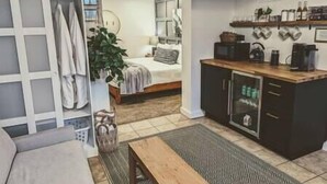 A compact living space with a kitchenette, a bed with a headboard, a dining area with a bench, and a large plant. - Hidden Garden Suite & Spa (Minneapolis)