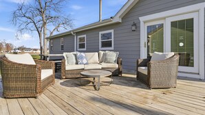 A covered deck with wicker furniture, a round table, and a gray house in the background. - Cozy Lakeside Cottage for Families and Fun Seekers. Game room - Large Soaker Tub (Council Bluffs)