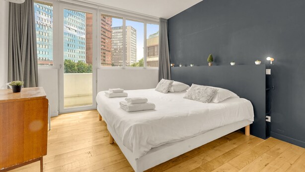 A modern bedroom with a large bed, a wooden dresser, a dark accent wall, and a view of buildings through the window. - Le Bonrepos - T3 sur les Ramblas de Toulouse (Toulouse)