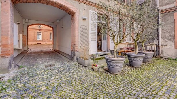 A cobblestone courtyard with a brick archway leading to a building entrance, surrounded by potted plants and outdoor furniture. - Bleu du Maine - Elegant T3 avec terrasse (Toulouse)