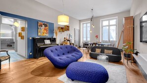 A modern living room with a piano, blue upholstered chairs, a dining area, and a large window. - Bleu du Maine - Elegant T3 avec terrasse (Toulouse)