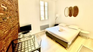 Un dormitorio moderno con pared de ladrillo, una cama con ropa de cama blanca, una mesita de noche y una pequeña mesita auxiliar redonda.