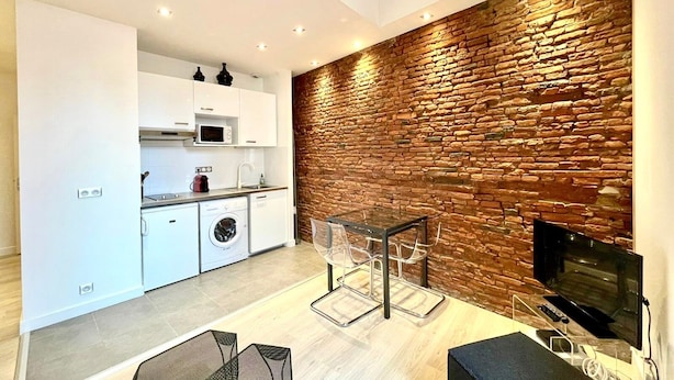 Una cocina moderna con armarios blancos, microondas y lavadora. Hay una mesa de vidrio con sillas y un televisor montado en una pared de ladrillo.